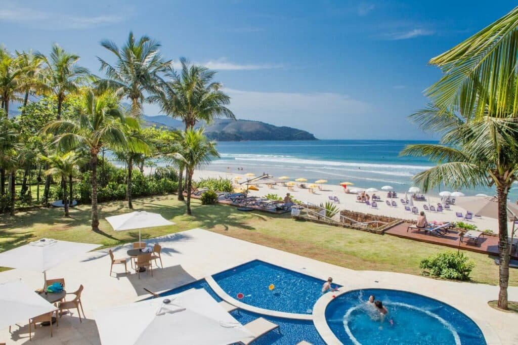 Piscina do Amora Hotel. Em volta dela estão algumas mesas de quatro lugares com guarda-sóis. Em frente há um gramado e espreguiçadeiras que ficam de frente para a praia.