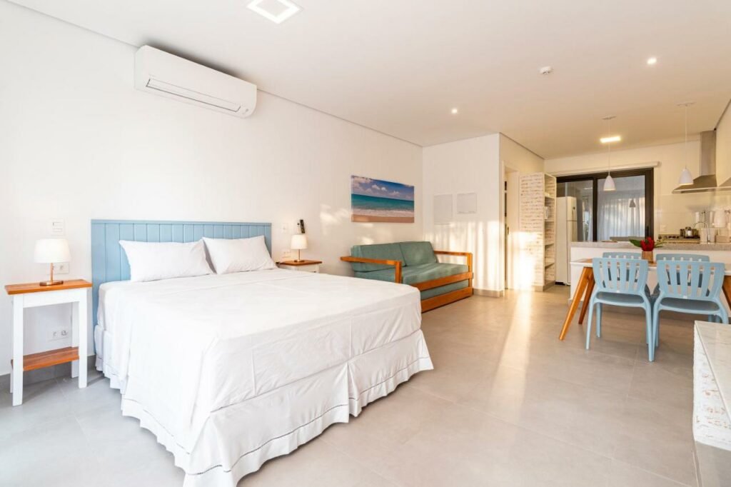 Quarto do Anoa Maresias Studios . Em primeiro plano está uma cama de casal com uma mesinha em cada lado. Perto dela e também encostado na parede está um sofá, de frente para uma mesa de quatro lugares. Ao fundo está a cozinha.