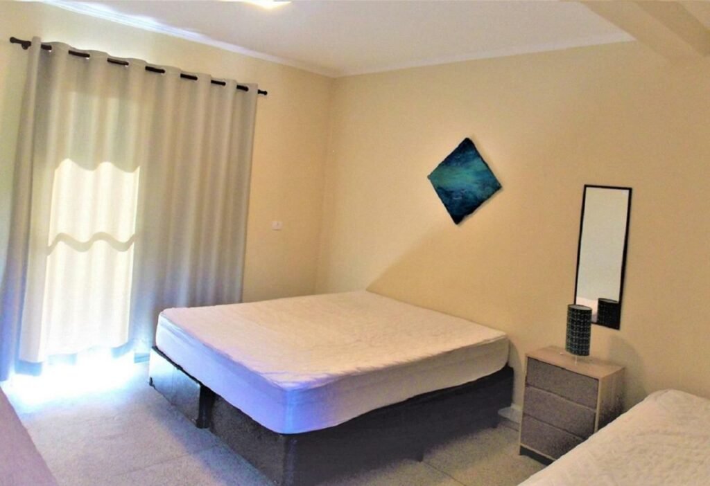 Quarto do "Apartamento com Vista pro Mar". Uma cama de casal está no meio, ao lado de uma mesinha com gavetas e outra cama. No canto há uma janela com cortinas.