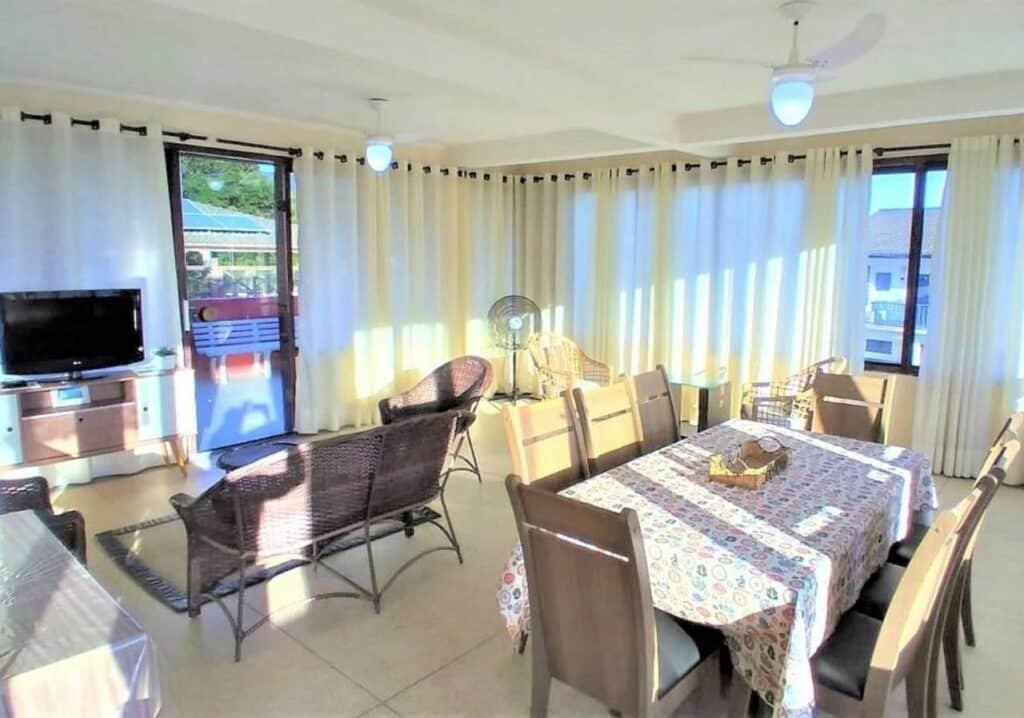 Sala de estar do "Apartamento com Vista pro Mar". Do lado esquerdo está a tv, perto das janelas e de frente para um sofá e uma poltrona. Do lado direito há uma mesa de vários lugares. O ambiente é todo cercado por janelas com cortinas.