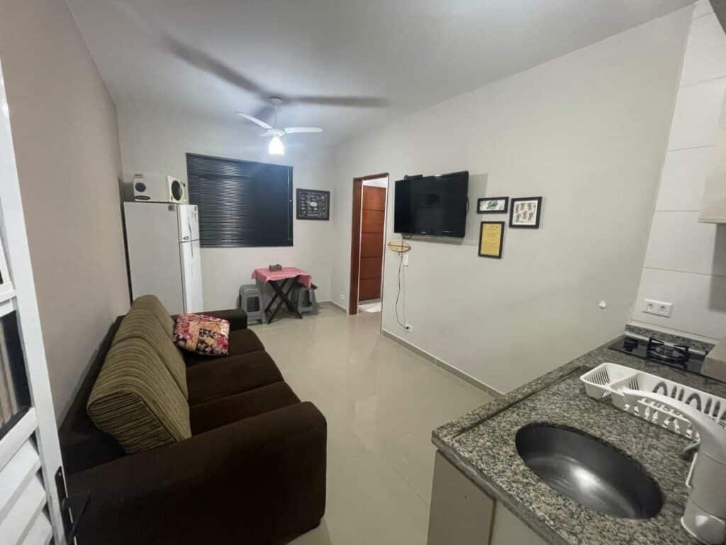 Área de estar e pequena cozinha do Apê 06 Maresias. Do lado direito estáa uma pia e uma boca de fogão. No meio e de frente para uma tv na parede está o sofá. Do lado esquerdo há uma geladeira com um micro-ondas em cima, uma mesinha com dois bancos e uma porta levando para outro cômodo.