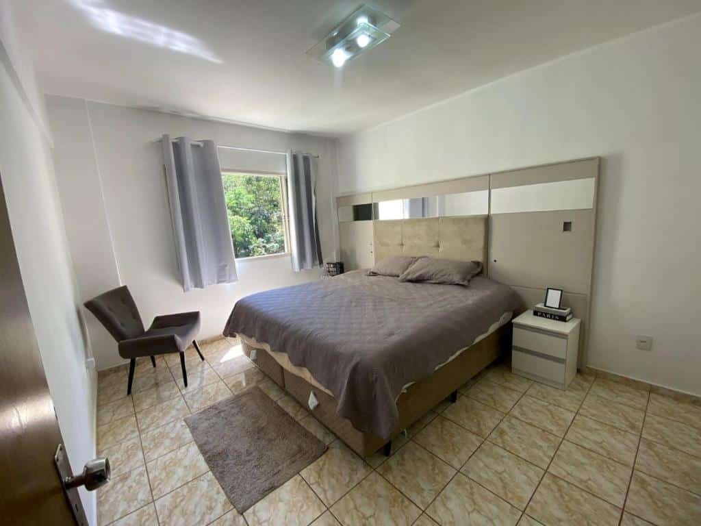 Foto do quarto em Apartamento VIP Centro Serra Negra. O quarto é grande e possui uma cama box de casal com cabeceira espelhada no centro. Na esquerda da cama há uma janela e uma poltrona preta.
Ilustra o post sobre airbnb em Serra Negra.