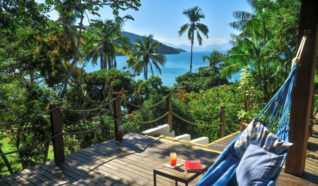 Imagem de uma das varandas da pousada Atlantica Jungle Lodge. No canto direito há uma rede de descanso e uma pequena mesinha com um livro em cima. Já no lado esquerdo há uma escada de madeira e uma vista para uma vegetação e o oceano ao fundo.