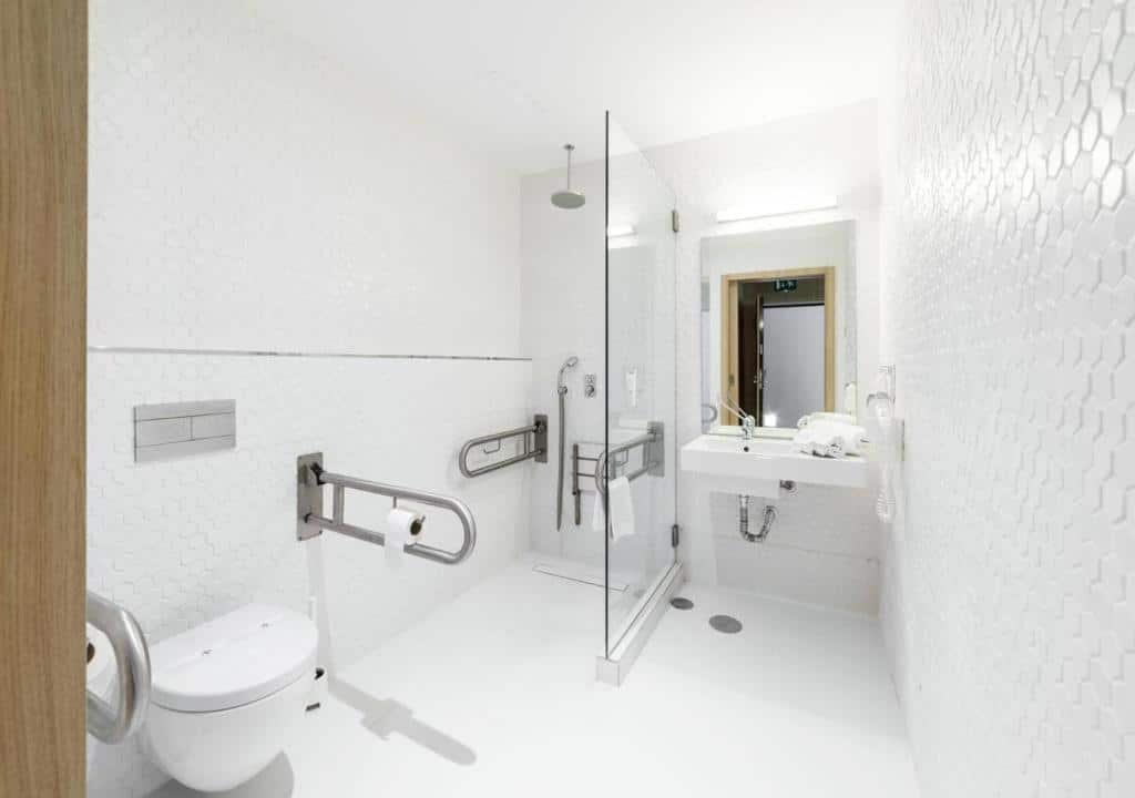 Banheiro com acessibilidade do Moov Hotel Porto Centro com vaso sanitário do lado esquerdo com barras de segurança, logo ao fundo o chuveiro e do lado direito pia baixa.