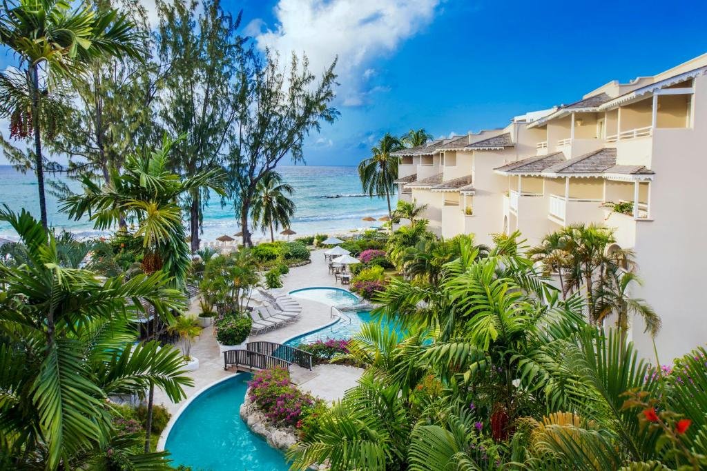Parte externa do Bougainvillea Barbados que mostra várias árvores verdes e algumas plantas coloridas. Na parte de baixo da imagem tem uma piscina em formato de rio, do lado direito tem a construção do hotel e ao fundo o mar. Imagem ilustrando post Hotéis em Barbados.