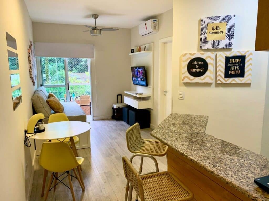 Imagem da sala de estar, balcão da cozinha e mesa de jantar do Boulevard Riviera Flat – Praia, tudo decorado, e uma sacada com vista para o jardim