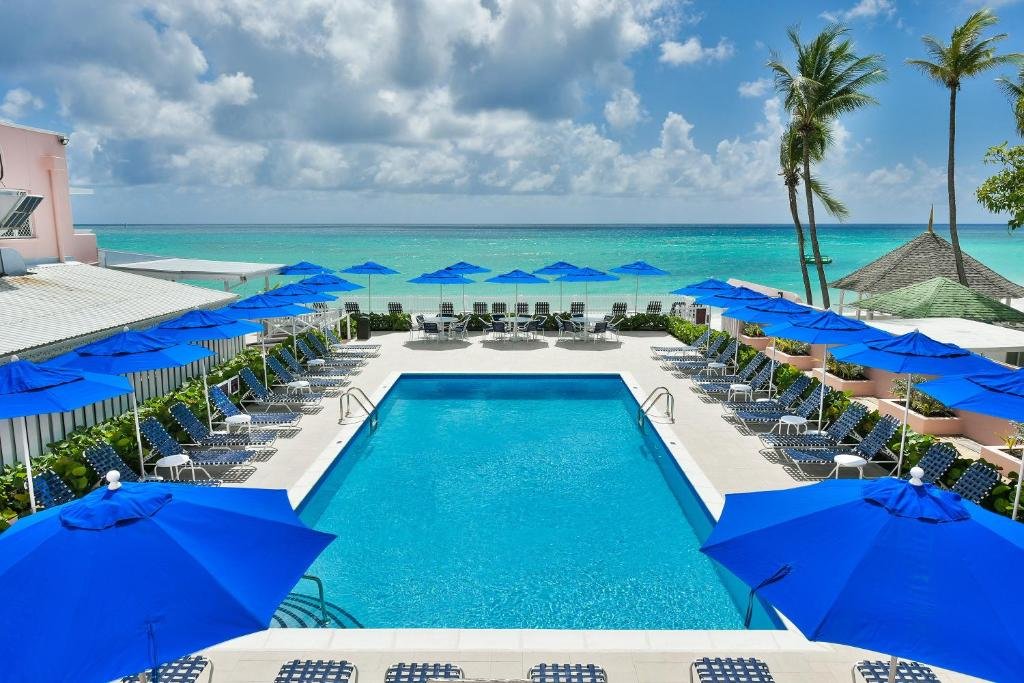 Área externa do Butterfly Beach Hotel, com uma piscina no centro, a redor cadeiras de tomar sol e guarda-sóis, no fundo o mar e palmeiras. Imagem ilustrando post Hotéis em Barbados.
