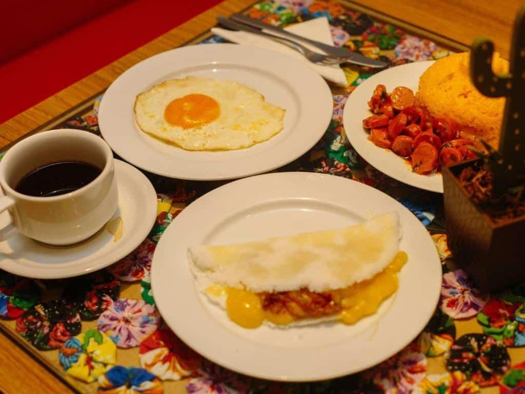 Pratos com tapioca, cuscuz, salsicha, ovo, café, talheres ao lado e tecido florido em baixo.