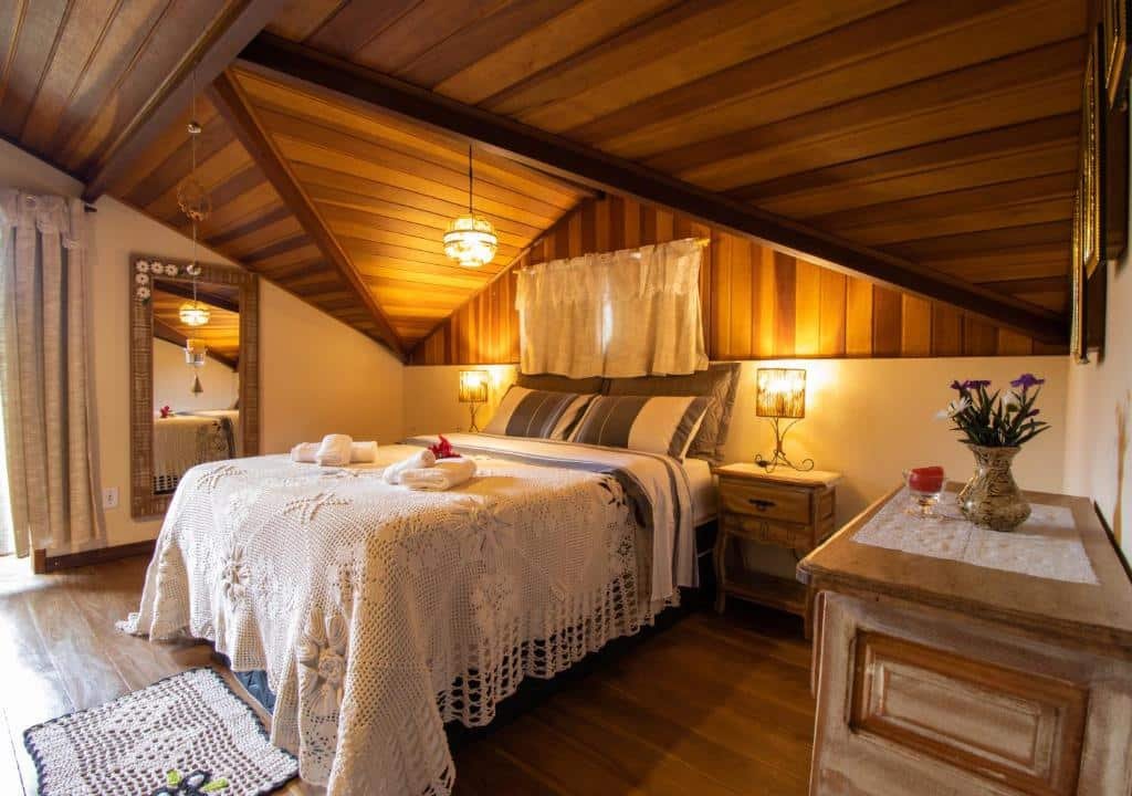 Imagem do quarto em Cantinho Pedra da Gávea, que está ilustrando o post sobre airbnb em Visconde de Mauá. O quarto é espaçoso, há uma cama box de casal no meio, e nos dois lados dela há uma mesa de cabeceira com abajur. No fundo à esquerda há um espelho e uma porta com varando, e no canto direito há uma cômoda.