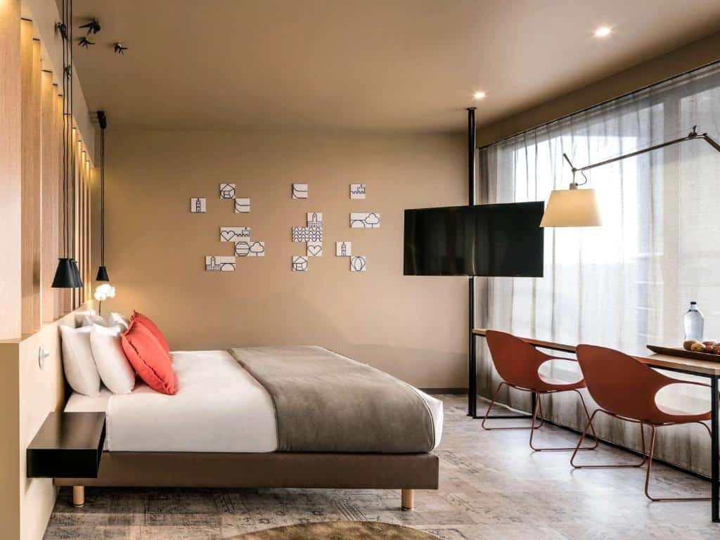Quarto do Mercure Porto Centro Santa Catarina com cama de casal do lado esquerdo da imagem, em frente a cama uma mesa com duas cadeiras e do lado esquerdo da cama em frente uma TV.