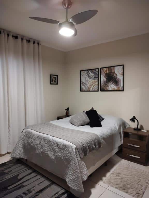 Foto do quarto em Casa agradável com churrasqueira no centro de Serra Negra. Há uma cama de casal ao centro. De seus dois lados há uma mesa de cabeceira de madeira com abajur em cima. Na parede atrás da cama há dois quadros decorativo. Na esquerda há uma janela com cortinas brancas. 
Ilustra o post sobre airbnb em Serra Negra.