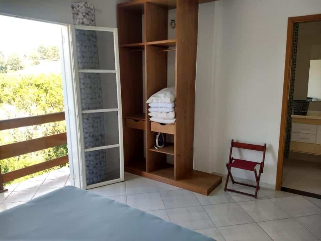 Foto do quarto em Casa com linda Vista no Centro. A cama está no canto inferior esquerdo vemos parte do pé do colchão. De frente há uma estante de madeira com almofadas, e de seu lado esquerdo há uma varanda. Na direita há uma porta para o banheiro. 
Ilustra o post sobre airbnb em Serra Negra.
