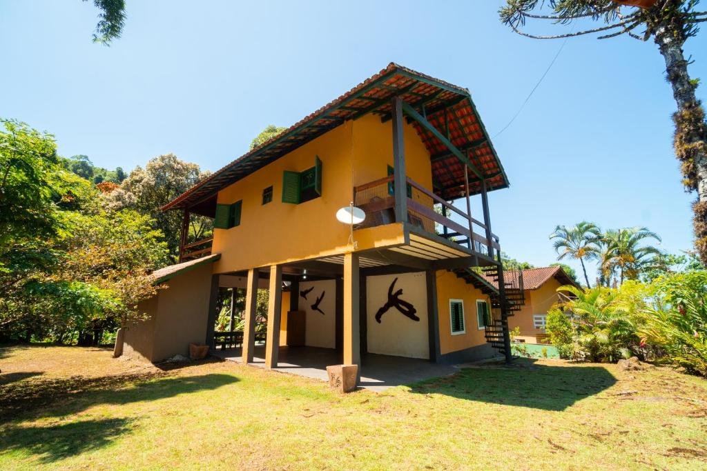 Fora da Casa da Almada. Um sobrado com escadas na área externa, uma varanda, e um jardim em baixo cercado de árvores.