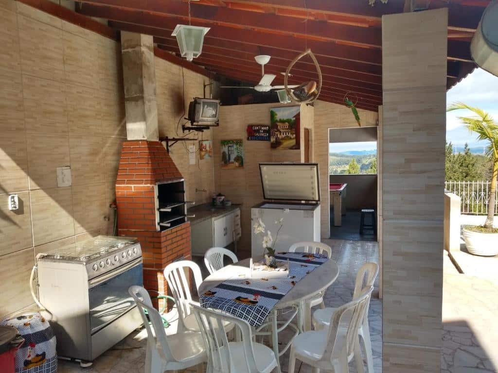 Foto da área de lazer em Chácara Recanto Paraíso. Há uma cozinha ao ar livre com mesa, cadeiras, fogão, churrasqueira, pia e freezer. Também há uma TV no topo de uma parede ao fundo. O rancho é coberto por um telhado. 
Ilustra o post sobre airbnb em Serra Negra.