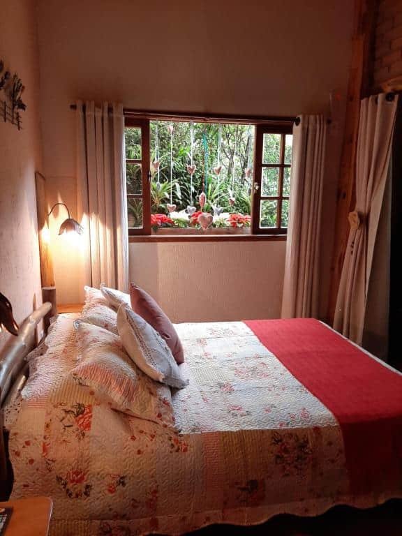 Imagem do quarto em Chalé Raio De Luz, que está ilustrando o post sobre airbnb em Visconde de Mauá. Vemos a cama de lado, e é de casal, e do outro lado dela há uma janela com decorações românticas e vista para um jardim.