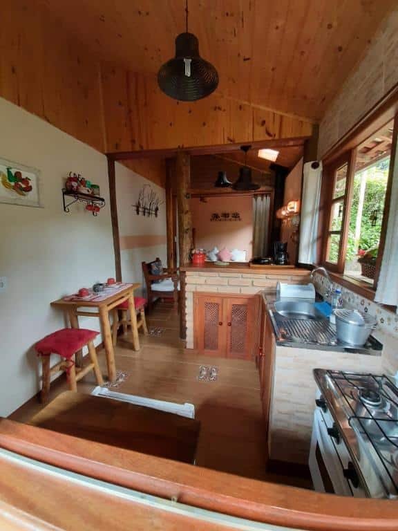 Foto mostrando a cozinha do Chalé Raio De Luz. Há um fogão pia, armários e mesa com dois bancos. Uma bancada separa esse cômodo da sala.