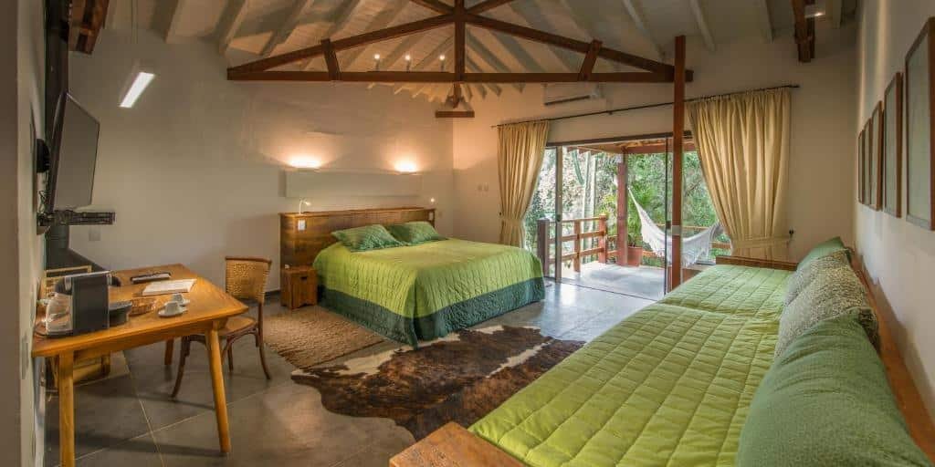 Quarto de chalé de luxo com cama de casal com colchas verdes, grande sofá com colcha verde, mesa e cadeira de madeira, TV, cortinas bege e varanda com rede branca. Imagem para ilustrar o post hotéis e pousadas em Itu.