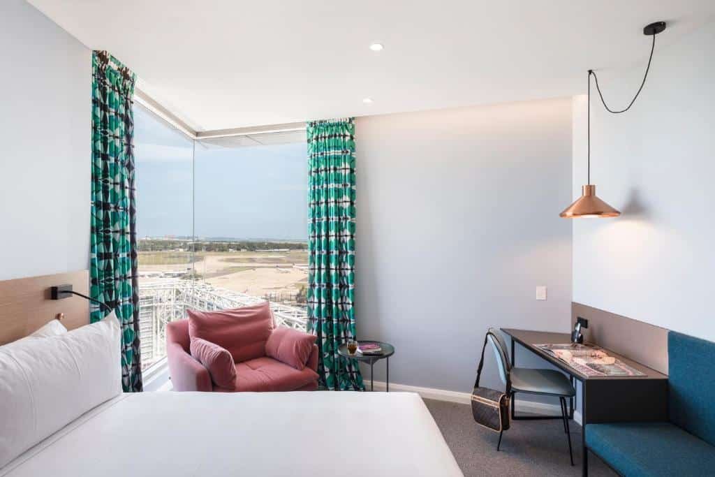 Quarto do Citadines Connect Sydney Airport. Uma cama de casal, uma poltrona e uma parede de vidro do lado esquerdo. Do lado direito uma mesa de trabalho e um sofá.  Imagem para ilustrar post sobre hotéis em Sydney.