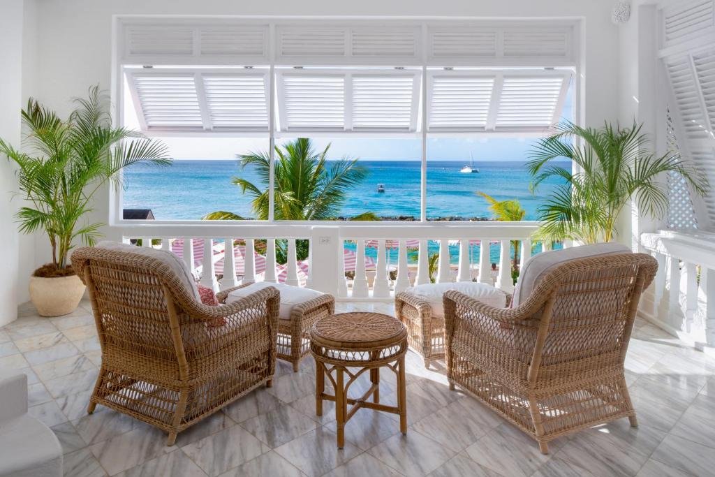 Parte do Cobblers Cove que mostra duas poltronas com dois bancos aos pés e uma mesinha redonda no meio delas. Essas poltronas estão em uma área fechada na cor branca, tem duas plantas verdes e uma parte aberta que tem vista para a paisagem com árvores e um mar azul. Imagem ilustrando post Hotéis em Barbados.