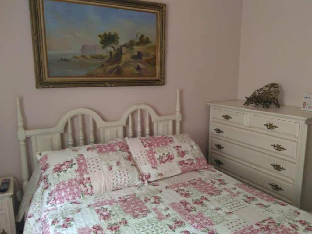 Foto do quarto em Condomínio Vila Paradiso. As paredes são rosa claro. Há uma cama no centro com cabeceira de madeira branca, e ao seu lado na direita há um gaveteiro branco de madeira. Há um quadro atrás da cama, decorando a parede. 
Ilustra o post sobre airbnb em Serra Negra.