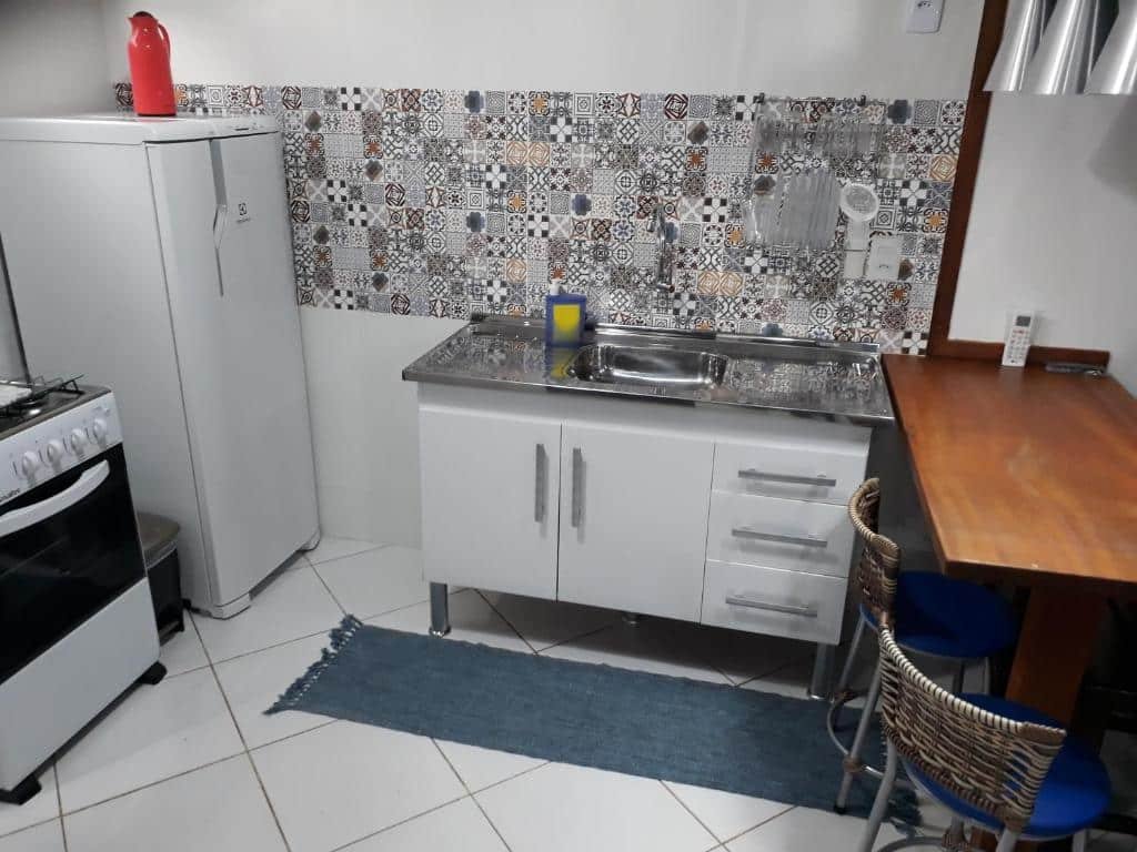 Cozinha do airbnb Loft e Vila da Praia Studios Ilha Grande. No lado direito há uma mesa com duas cadeiras, no meio há um armário baixo com a pia e no canto esquerdo há uma geladeira e o fogão.