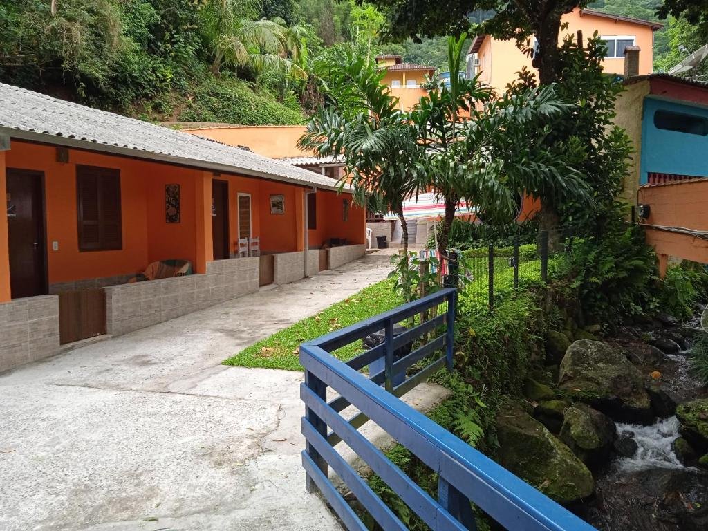Entrada da Pousada Caiçara, construção em tons de laranja, uma corrimão azul protege um pequeno lago com pedras, muitas árvores e um gramado