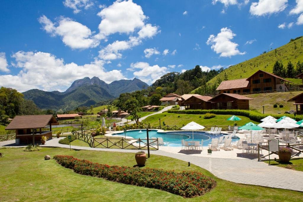 A Fazenda Suíça Le Canton conta com piscina, espreguiçadeiras e chalés de madeira cercados por colinas verdes sob um céu azul.