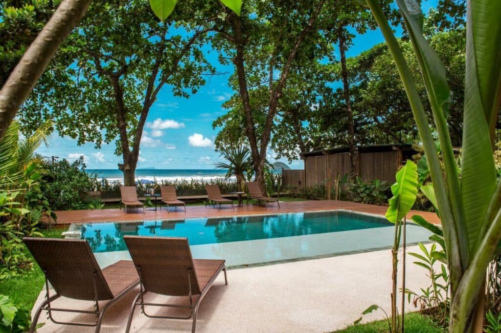 Espreguiçadeiras encarando a piscina cercada por plantas no Flat Pitanga, um dos airbnb em Camburi. Ao fundo é possível ver um pouco da praia. O céu acima é azul com poucas nuvens.