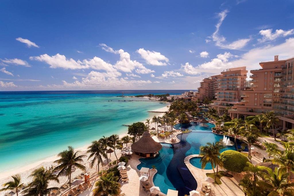 vista da sacada do Grand Fiesta Americana Coral Beach Resort Cancun mostrando a praia de area clara, o mar de tons turquesa e o céu azul. À direita é possível ver a piscina do resort e alguns prédios em tons rosado