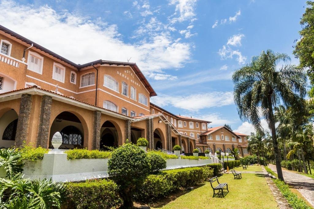 Exterior do Grand Resort Serra Negra. O céu está azul com nuvens, há um jardim na entrada com árvores, gramado e arbusto e o hotel está logo atrás.