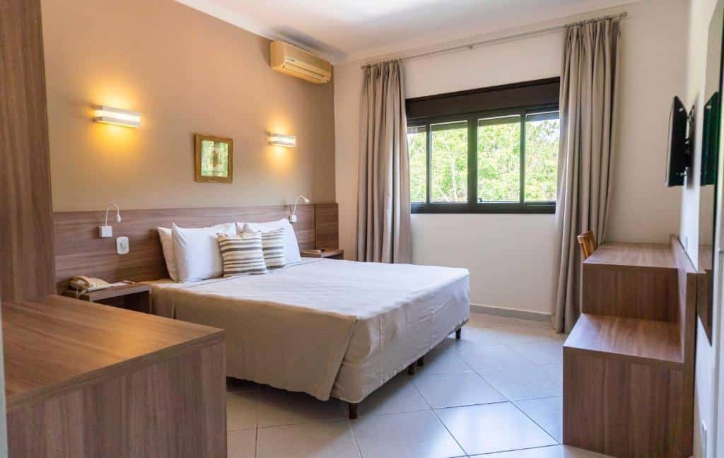 Quarto Triplo Standard do Guararema Parque Hotel, de 21 m², com cama de casal, ar-condicionado, TV suspensa na parede e móveis de madeira. Tem uma janela no lado direito com vista da natureza