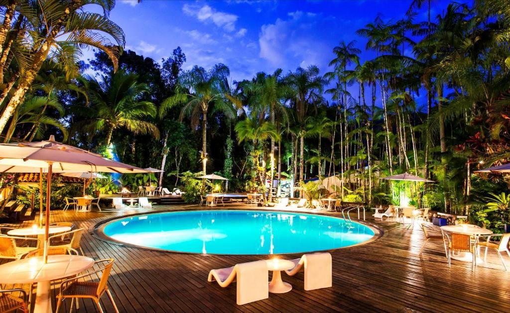 Piscina em Hotel Aldeia de Sahy, uma piscina redonda cercada por um deck, muitas árvores, uma iluminação agradável de noite, muitas cadeiras e mesas com guarda-sóis ao redor. Representa airbnb na Barra do Sahy.