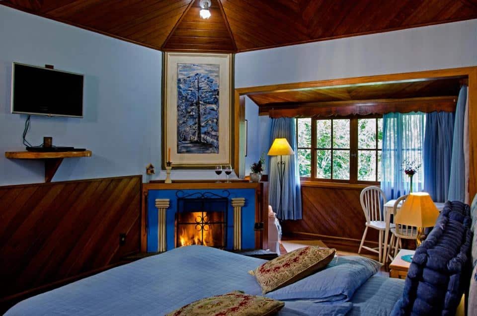 Quarto no Hotel Buhler em Visconde de Mauá. As paredes são azuis e todos os outros detalhes do quarto também, misturado com madeira escura. A cama está no canto direito. Na frente dela há uma lareira e uma televisão. Ao seu lado, no fundo do quarto há um abajur, uma mesa com cadeiras e uma janela.