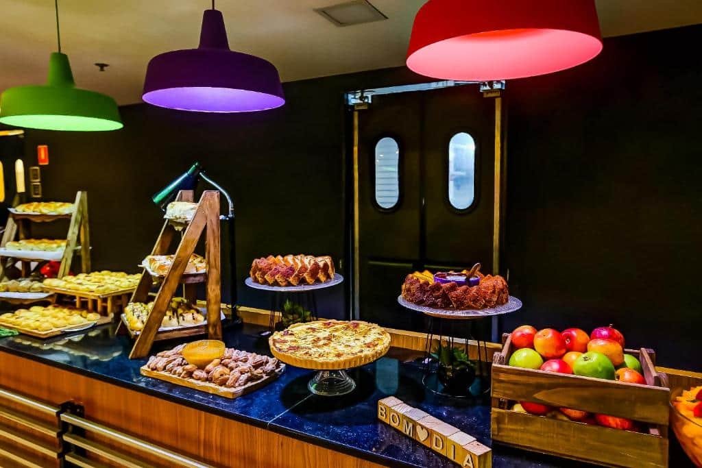 Bancada com café da manhã com frutas, bolos e outras comidas expostas e lustres coloridos.