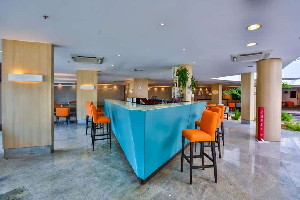Balcão azul com cadeiras laranjas, pilastras com detalhes em madeiras e algumas plantas verdes na parte externa do hotel.