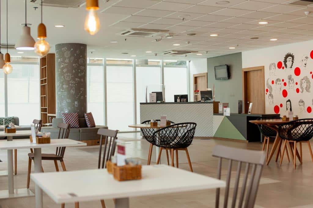 Recepção do hotel com algumas mesas e cadeiras espalhadas, detalhes verdes na parede e alguns desenhos pretos e vermelhos na parede branca.