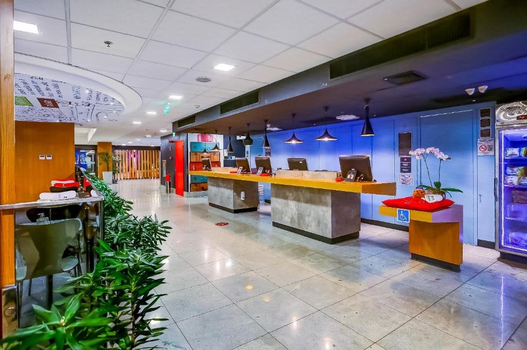 Imagem da parte interna do hotel com bancadas para atendimento, algumas mesas com cadeiras e uma planta verde.
