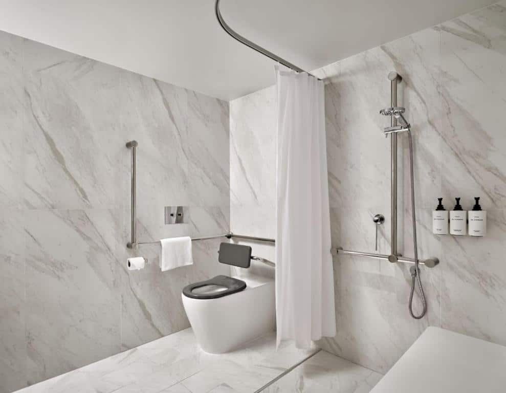 Banheiro do InterContinental Sydney, an IHG Hotel. Do lado direito um box, cadeiras e barras. Do lado esquerdo um vaso sanitário e barras de apoio.