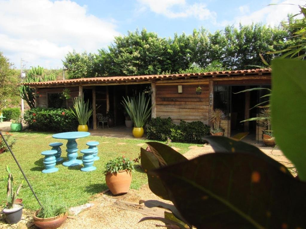 Exterior do La Villa Chalés Garden. Vemos um rancho de madeira, com muitos vasos e arbustos. Há um gramado à frente com mais plantas e uma mesa de pedra com bancos pintados de azul no meio.