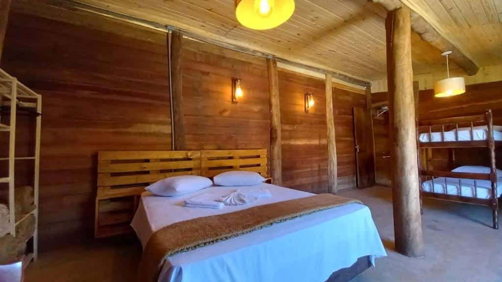 Foto do quarto em La Villa Chalés Garden. O chalé tem paredes de madeira. Há uma cama de casal no meio, uma estante à sua esquerda e na direita há uma beliche.  
Ilustra o post sobre airbnb em Serra Negra.