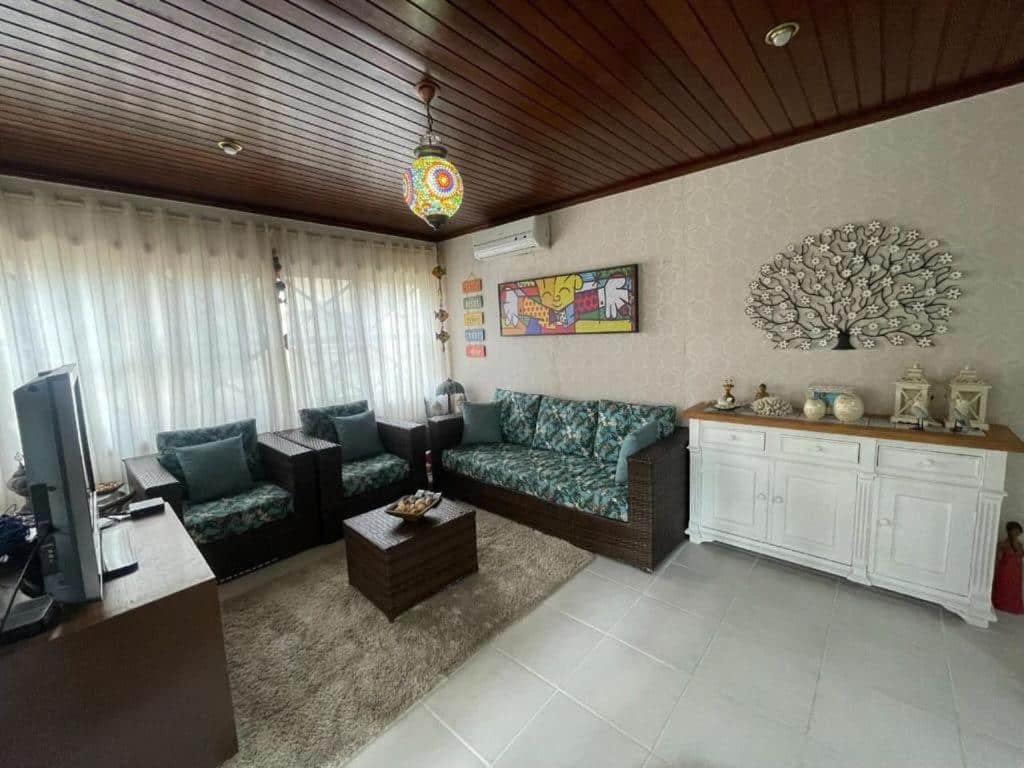 Sala de estar do airbnb "Linda Casa Condomínio Maresias". Do lado esquerdo, perto de uma grande janela com cortinas, estão duas poltronas e um sofá. Em frente a eles está uma tv e uma mesinha no meio. Do lado direito está um móvel com aguns armários e decorações