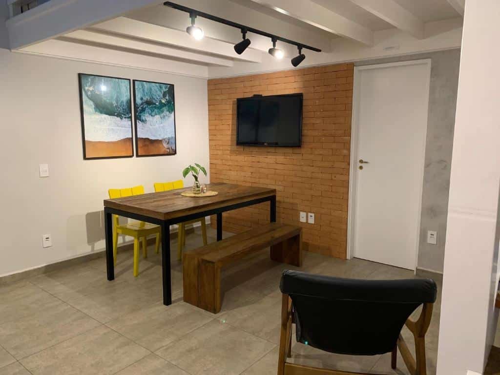Área de estar do "Lindo Flat em Maresias, 100m da Praia". De um lado, perto da parede, está uma mesa de vários lugares. De frente para ela há uma tv. Ao lado da tv está uma porta. Do outro lado há uma cadeira.