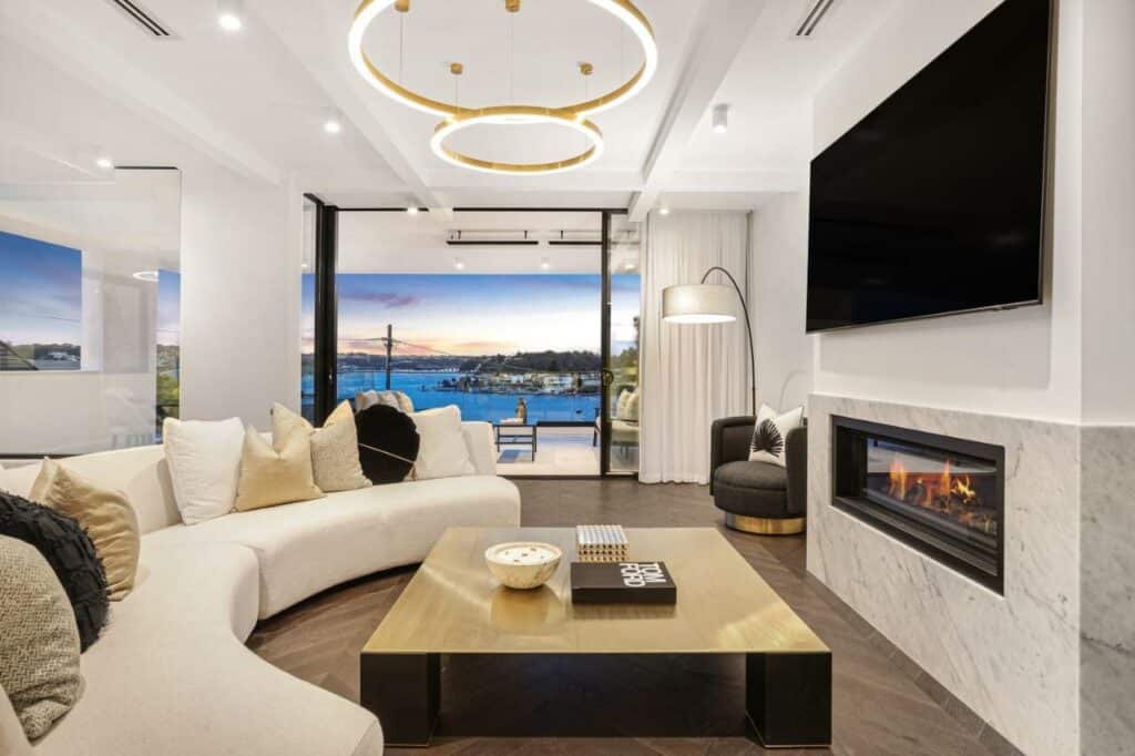 Sala do Luxury Waterside Home. Um sofá do lado esquerdo, no centro ua mesa e em cima um lustre. Do lado direito uma lareira, uma TV, um abajur com uma poltrona. No fundo a varanda da casa.