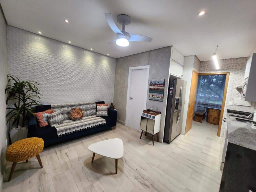 Sala de estar e cozinha do Maresias Beach Home - Mata Azul. Do lado esquerdo está a sala, com um sofá, um banquinho e uma mesa no meio. Há também um ventilador de teto e uma porta fechada ao lado do sofá. Do lado direito está a cozinha, com uma pia encostada na parede, armários na parede e uma geladeira na parede oposta.