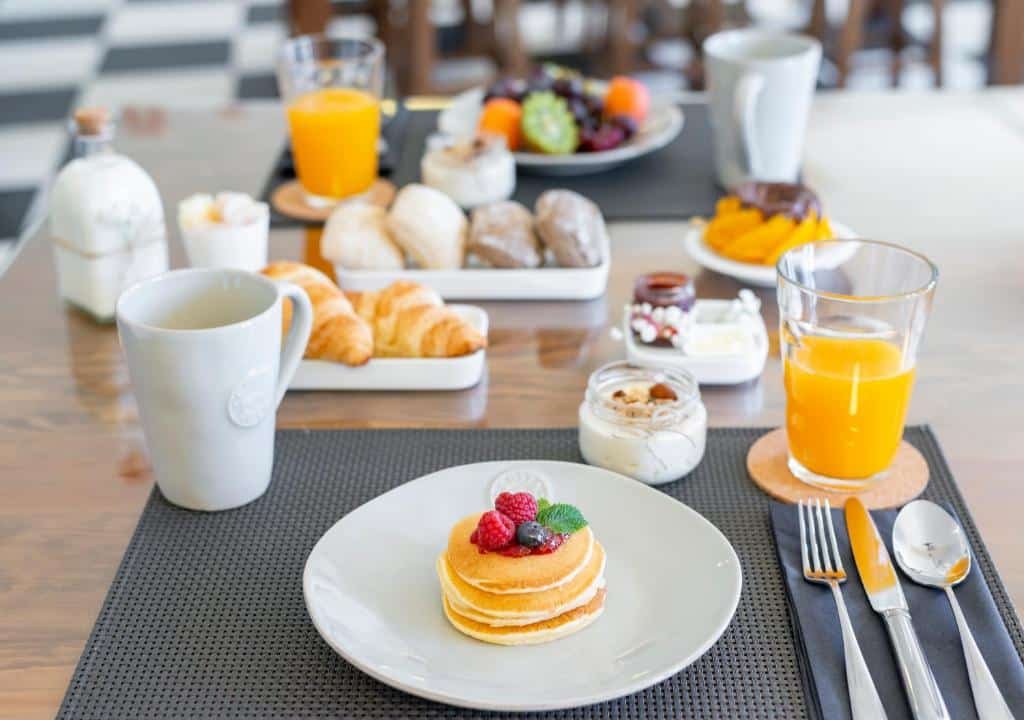 Mesa de café do Oporto Street Fonte Taurina - Riverfront Suites com um prato com uma pequena panqueca com frutas em cima, do lado direito um copo com suco e do lado esquerdo uma xícara e ao fundo vários pães e frutas.