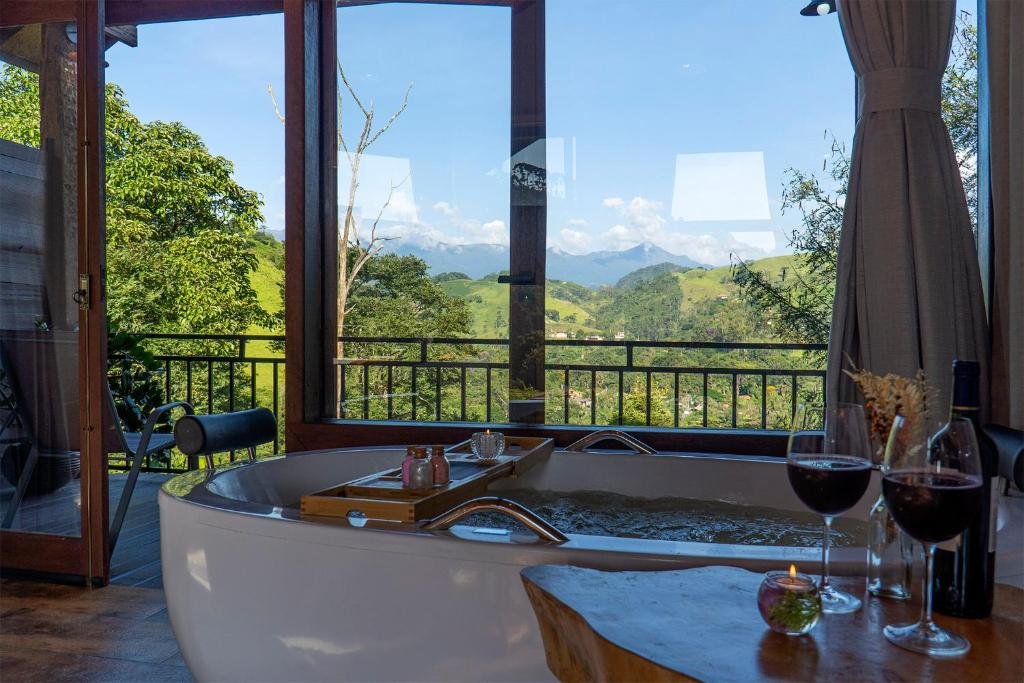 Monte Mauá Chalés. Vemos a banheira de perto, e nela há amenidades para banho em cima de uma superfície. No canto inferior direito há duas taças de vinho. A banheira está de frente para uma varanda com vista.