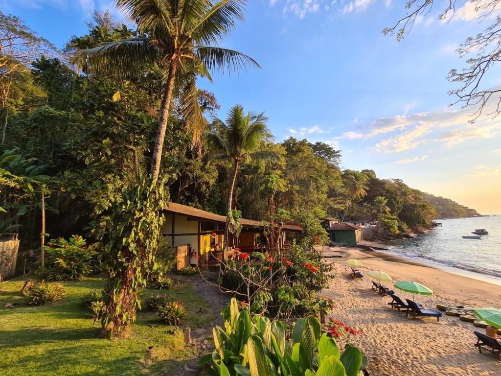 Hotel Paraíso Azul Retiro visto do lado de fora. O hotel fica na beira de uma praia, atrás dele há muita vegetação e na sua frente está o mar e algumas cadeiras de praia na areia. Imagem para ilustrar o post airbnb em Ilha Grande.