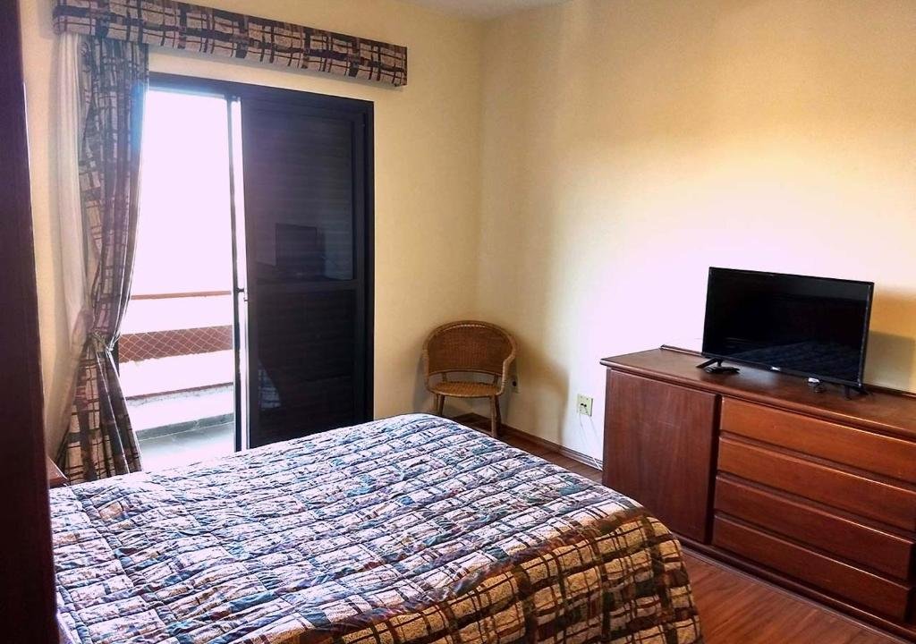 Foto do quarto em Piemonte Flat Serra Negra. A cama está no meio, vemos ela de lado. Na frente dela há uma estante de madeira com uma pequena TV em cima. Do outro lado do quarto há uma porta com varanda.  
Ilustra o post sobre airbnb em Serra Negra.