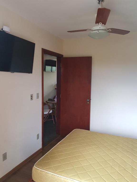 Foto do quarto em Piemonte Flat. Há uma cama de casal no canto inferior direito, vemos o colchão de relance. De frente para a cama há uma TV fixa na parede. No teto há um ventilador.  porta também está de frente com a cama. 
Ilustra o post sobre airbnb em Serra Negra.