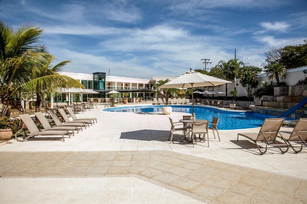 Piscina azul com espreguiçadeiras bege em volta, guarda-sol, palmeiras e hotel de paredes brancas ao fundo.